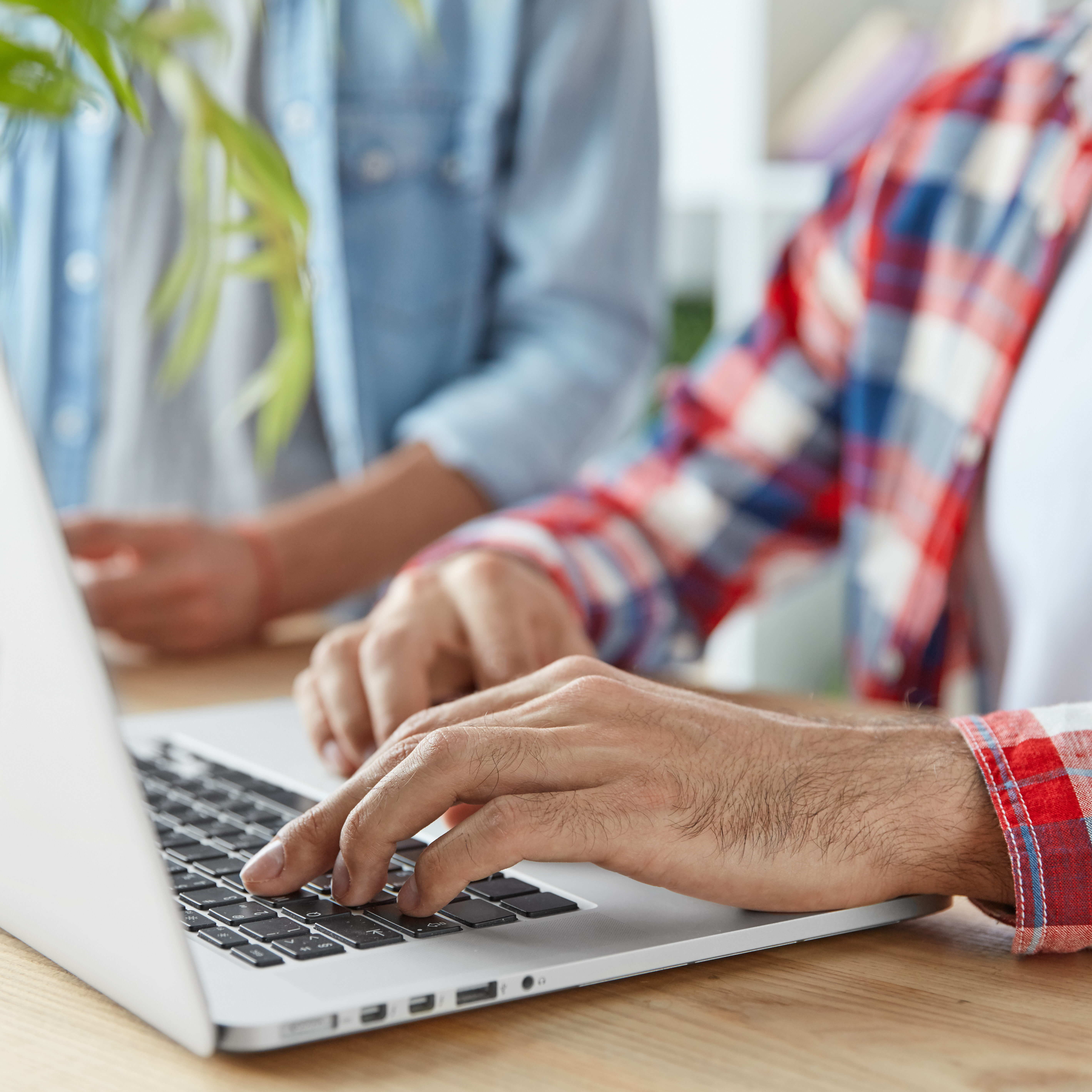 Cropped shot of two male bloggers type publication on laptop computer, use laptop computer, sit at wooden desk. Young prosperous bussinessmen check mail and send feedbacks, connected to wifi