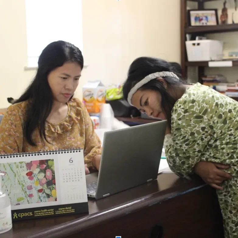 one woman sitting at a desk in front of a computer with another woman leaning over the desk to look at the screen